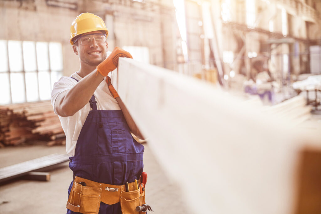Smiling man builder carrying wooden plank at construction site
