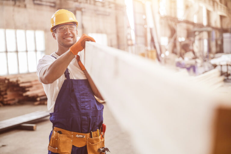 Smiling man builder carrying wooden plank at construction site